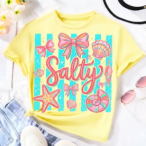May include: A pale yellow short-sleeve t-shirt featuring a central graphic with the word Salty in pink script. The design includes vertical turquoise stripes, pink bows, a starfish, various seashells, and small fish illustrations in a colorful palette.