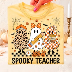 May include: A light yellow t-shirt featuring a graphic of three ghosts. One ghost has a leopard print pattern and a book on its head, one wears a polka dot bow and holds a pencil, and one has a checkered pattern and holds a stack of books. The text reads SPOOKY TEACHER.