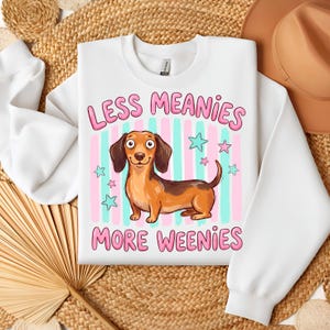 Puede incluir: Sudadera blanca de cuello redondo con un dibujo de un perro salchicha de dibujos animados. El diseño incluye el texto "LESS MEANIES MORE WEENIES" en rosa, con un fondo de rayas rosas y azules y estrellas verdes.