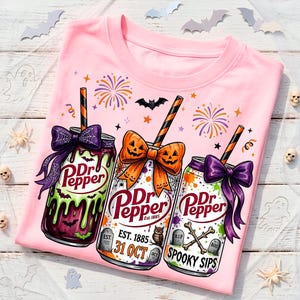May include: A light pink t-shirt featuring a graphic of three Dr Pepper soda cans decorated for Halloween. The cans are adorned with bows, striped straws, and spooky motifs like tombstones, bats, and dripping green liquid with text.