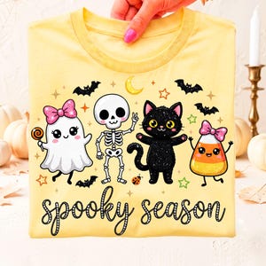 May include: A pale yellow t-shirt featuring a graphic print of a ghost with a pink bow, a skeleton, a black cat, and a piece of candy corn with a pink bow. The text "spooky season" is printed in black cursive lettering below the characters.