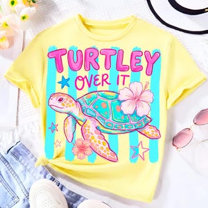 May include: A bright yellow short-sleeve t-shirt featuring a graphic of a sea turtle, pink hibiscus flowers, and blue stars. The text "TURTLEY OVER IT" is printed in bold pink letters above the turtle against a background of vertical blue stripes.