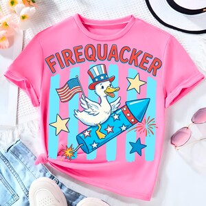 May include: Pink t-shirt with the word "FIREQUACKER" in red letters. The shirt features a cartoon duck wearing a patriotic hat and holding an American flag, riding a blue rocket with stars and fireworks.