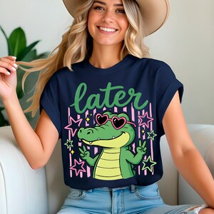 May include: Navy blue t-shirt featuring a cartoon alligator wearing heart-shaped sunglasses and making a peace sign. The word "later" is written in green script above the alligator. Pink stars and stripes are in the background.