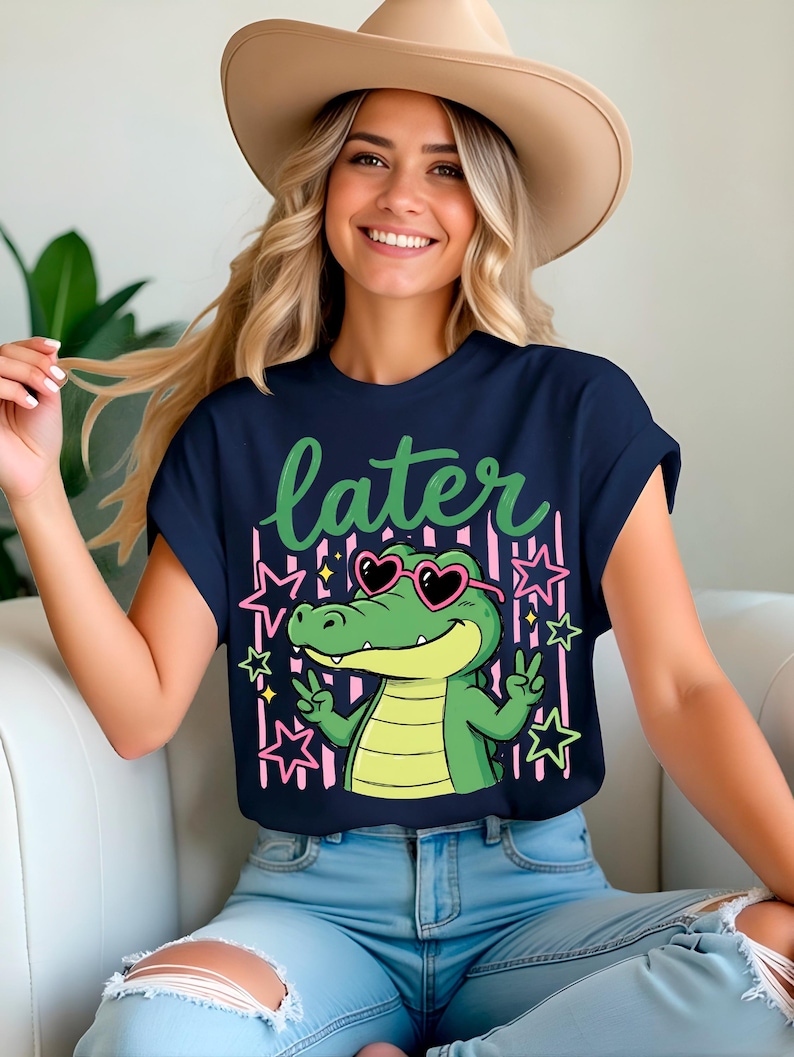 May include: Navy blue t-shirt featuring a cartoon alligator wearing heart-shaped sunglasses and making a peace sign. The word "later" is written in green script above the alligator. Pink stars and stripes are in the background.