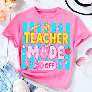 May include: A pink t-shirt with the words "TEACHER MODE" in yellow and pink, accented with a pencil, flowers, stars, and an apple. The shirt has a power button graphic that says "OFF". The shirt is short-sleeved.