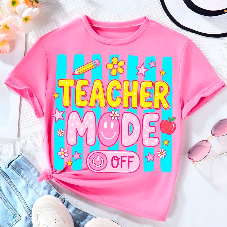 May include: A pink t-shirt with the words "TEACHER MODE" in yellow and pink, accented with a pencil, flowers, stars, and an apple. The shirt has a power button graphic that says "OFF". The shirt is short-sleeved.