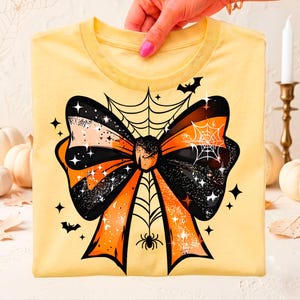 May include: A pale yellow t-shirt featuring a large graphic of a bow colored in black and orange. The bow design includes a spider web pattern, a small spider, several four-pointed stars, and two silhouettes of flying bats against the fabric.