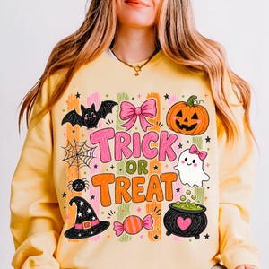 May include: A pale yellow long-sleeve sweatshirt featuring a colourful Halloween graphic with the text TRICK OR TREAT. The design includes a black bat, a pink bow, an orange pumpkin, a spider web, a white ghost, a witch hat, and a black cauldron.
