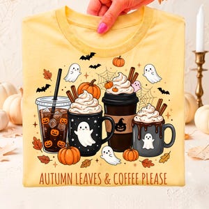 Peut inclure: Un t-shirt jaune pâle avec une impression graphique représentant trois boissons au café, des citrouilles, des fantômes, des chauves-souris et des toiles d'araignée. Le texte "AUTUMN LEAVES & COFFEE PLEASE" est imprimé en bas du motif.