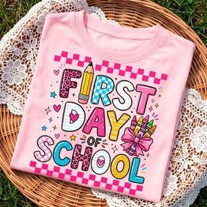 May include: A light pink t-shirt featuring a graphic print with the text FIRST DAY OF SCHOOL. The design includes a yellow pencil, a bundle of colorful crayons with a pink bow, and various patterns like leopard print, checkers, and hearts.
