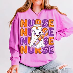May include: A light pink long-sleeve sweatshirt featuring a repeating orange and purple text pattern that reads NURSE. In the center, a white ghost character wears a nurse cap and holds a large orange syringe with a purple heart detail.