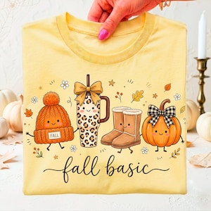May include: A pale yellow t-shirt featuring a graphic print of a smiling orange knit beanie with a 'FALL' tag, a leopard print tumbler with a bow, tan boots, and a pumpkin with a plaid bow, with the text 'fall basic' written in black script.