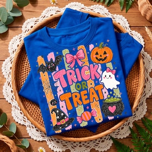 May include: A royal blue t-shirt featuring a colourful Halloween graphic with the text "TRICK OR TREAT". The design includes a pumpkin, a ghost, a witch hat, a bat, a spider web, a spider, a pink bow, and a cauldron filled with green potion.