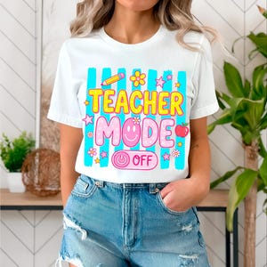 May include: White t-shirt with a colorful graphic that reads "TEACHER MODE OFF." The design includes a pencil, flowers, stars, an apple, and a smiley face. The shirt has a crew neck and short sleeves.