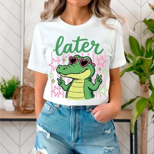 May include: White t-shirt with a cartoon alligator wearing heart-shaped sunglasses and making peace signs. The word "later" is written in green above the alligator. Pink and yellow stars and stripes are in the background.