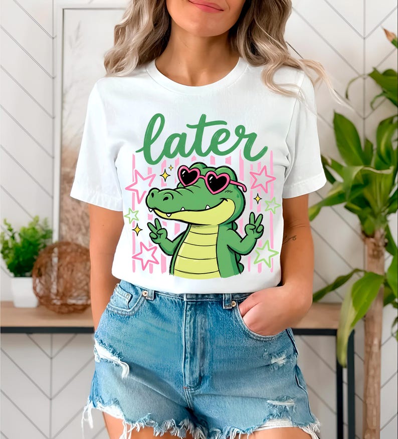May include: White t-shirt with a cartoon alligator wearing heart-shaped sunglasses and making peace signs. The word "later" is written in green above the alligator. Pink and yellow stars and stripes are in the background.