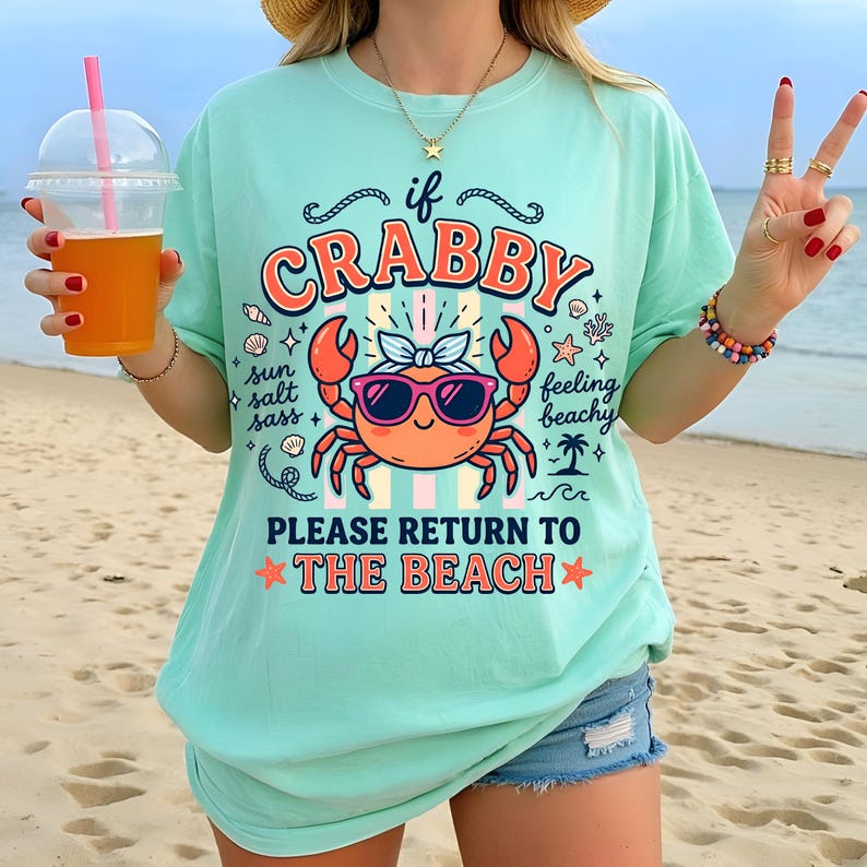 May include: A light teal t-shirt with a crab graphic wearing sunglasses and a bow. The text on the shirt reads "If Crabby, Please Return to the Beach." The shirt is paired with denim shorts.