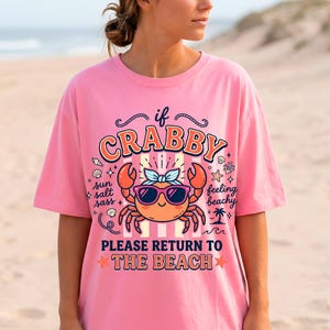 May include: A pink t-shirt featuring a cartoon crab wearing sunglasses and a bow. The text on the shirt says "if crabby, please return to the beach." The design includes stars, shells, and a palm tree.