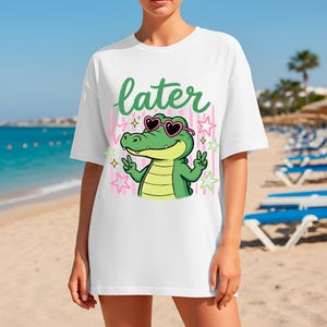 May include: White t-shirt featuring a cartoon alligator wearing heart-shaped sunglasses and giving a peace sign. The word "later" is written above the alligator in green script. Pink and white striped background with stars.