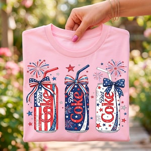 Puede incluir: Una camiseta rosa claro con un gráfico de tres latas de Diet Coke decoradas con motivos patrióticos en rojo, blanco y azul, incluyendo estrellas, rayas, fuegos artificiales y lazos, con cada lata rematada con una pajita y una cinta festiva.