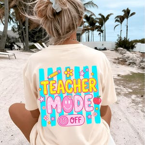 May include: Cream-colored t-shirt with a colorful graphic on the back that reads "TEACHER MODE OFF." The design includes a pencil, flowers, stars, a smiling face, and an apple. The shirt is worn on a beach.
