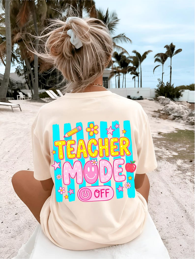 May include: Cream-colored t-shirt with a colorful graphic on the back that reads "TEACHER MODE OFF." The design includes a pencil, flowers, stars, a smiling face, and an apple. The shirt is worn on a beach.