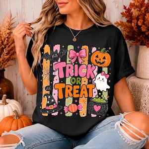 May include: A black t-shirt featuring a colourful Halloween graphic with the text "TRICK OR TREAT" in pink and orange. The design includes a pumpkin, a ghost, a witch hat, a bat, a spider, a bow, and a cauldron against vertical stripes.