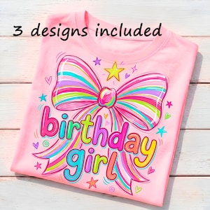 May include: A light pink t-shirt featuring a large, colorful striped bow graphic with the text "3 designs included" at the top and "birthday girl" in multi-colored lettering below the bow, surrounded by small stars and heart icons.