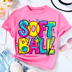May include: A pink t-shirt with the word "SOFTBALL" in a colorful, cartoonish design. The letters are outlined in blue and yellow, with a baseball and bat graphic. The shirt is displayed with denim shorts and white sneakers.