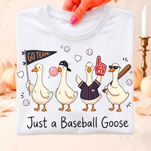 May include: A white t-shirt featuring a graphic of four cartoon geese in baseball gear. One holds a 'GO TEAM!' pennant, one blows a pink bubble, one wears a jersey with a #1 foam finger, and one holds a bat. The text 'Just a Baseball Goose' is below.