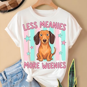 Puede incluir: Camiseta blanca con el texto rosa "LESS MEANIES MORE WEENIES". La camiseta presenta un perro salchicha de dibujos animados con orejas marrones y cuerpo marrón claro. Rayas y estrellas rosas y azules están detrás del perro.