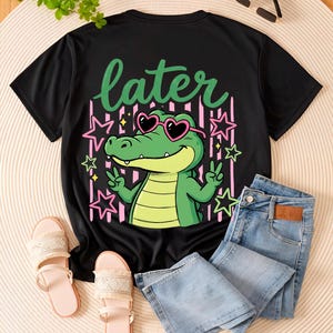 May include: Black t-shirt with a cartoon alligator wearing heart-shaped sunglasses and the word "later". The alligator is green and the sunglasses are pink. The shirt is paired with light wash denim jeans and pink sandals.