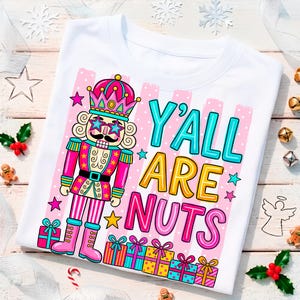 May include: A white t-shirt featuring a graphic of a nutcracker wearing star-shaped sunglasses, a crown, and a pink and gold uniform. The text Y'ALL ARE NUTS is printed in large, colourful letters next to the nutcracker, with gift boxes below.