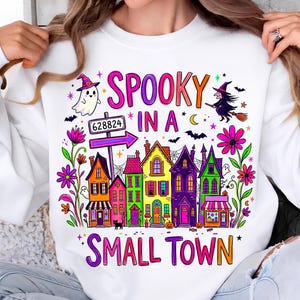 May include: A white sweatshirt featuring a colorful graphic of a small town with houses in shades of orange, pink, green, and purple. The text reads "SPOOKY IN A SMALL TOWN" with illustrations of a ghost, a witch on a broom, bats, and flowers.