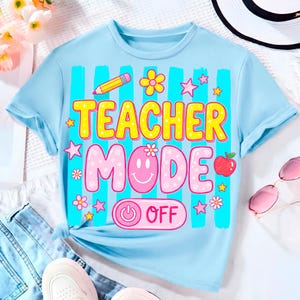 May include: Light blue t-shirt with the words "TEACHER MODE OFF" in a colorful design. The shirt features a pencil, flowers, stars, and an apple graphic. The shirt is made of a soft material and is suitable for casual wear.