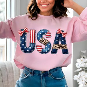 Może przedstawiać: Jasnoróżowa bluza z napisem "USA" w patchworkowym wzorze. Litery wykonane są z dżinsu, zwierzęcego nadruku i czerwono-białej tkaniny w paski, z czerwono-białymi kokardkami.