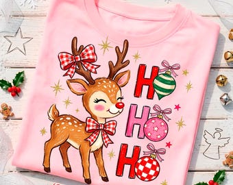 May include: A light pink t-shirt featuring a brown reindeer with a red nose, wearing a red and white checkered bow on its head and neck. The text HO HO HO is printed in red, accompanied by three hanging Christmas ornaments and gold stars.
