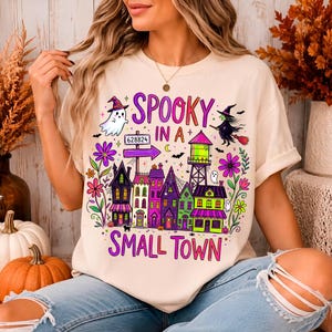 May include: A cream-colored t-shirt featuring a colorful graphic print with the text SPOOKY IN A SMALL TOWN. The design includes a ghost, a witch on a broomstick, a water tower, and a row of houses in purple, green, and pink, accented by flowers.