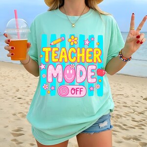 May include: A light green t-shirt with the words "TEACHER MODE OFF" in yellow and pink, decorated with a pencil, flowers, stars, and an apple. The shirt has a relaxed fit and is worn with denim shorts.