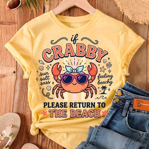 May include: A yellow t-shirt featuring a cartoon crab wearing sunglasses and a bow, with the text "If Crabby, Please Return to the Beach." The image includes blue jeans and beach accessories, creating a summery vibe.
