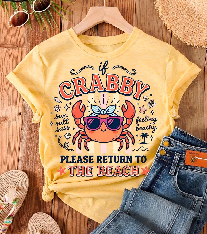 May include: A yellow t-shirt featuring a cartoon crab wearing sunglasses and a bow, with the text "If Crabby, Please Return to the Beach." The image includes blue jeans and beach accessories, creating a summery vibe.