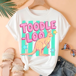 Op de afbeelding: Wit T-shirt met de tekst "TOODLE LOO" in roze letters, een cartoon kangoeroe en sterrenafbeeldingen. Het shirt wordt gestyled met sandalen en een spijkerbroek.
