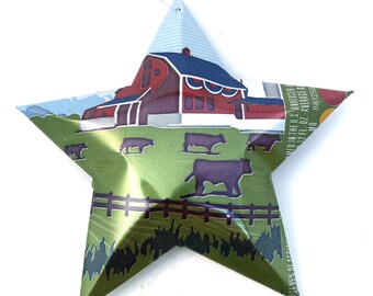May include: A five-pointed star ornament crafted from recycled aluminum, featuring a printed rural scene with a red barn, green pasture, and dark silhouettes of grazing cattle. A thin silver string is attached to the top point for hanging.
