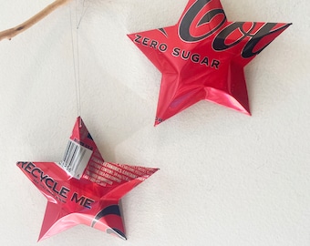 May include: Two red star-shaped ornaments made from recycled aluminum cans. The top ornament has the text "ZERO SUGAR" printed on it. The bottom ornament has the text "RECYCLE ME" printed on it.