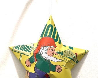 May include: A star-shaped ornament made from a yellow material, featuring a cartoon gnome running. The gnome wears a red hat, green shirt, and blue pants. The ornament is suspended by a string and is hanging from a small branch.