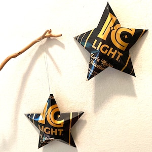 May include: Two star-shaped ornaments made from recycled beer cans. The stars are black and gold with the words "IC LIGHT" and "Let's Raise Another" printed on them. One star hangs from a thin wire attached to a small branch.