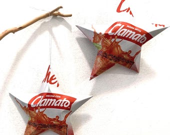 May include: Two star-shaped ornaments crafted from recycled Clamato packaging. Each star features the red and white Clamato logo, a graphic of a tomato juice splash, and a dark rim. One star hangs from a thin silver string attached to a wooden branch.