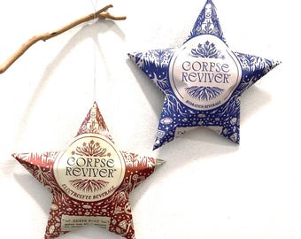 May include: Two star-shaped beverage pouches hanging from a wooden branch. The left pouch is red with the text Corpse Reviver Electrolyte Beverage Guava Rose. The right pouch is blue with the text Corpse Reviver Hydration Beverage. Both feature intricate botanical and crystal illustrations.