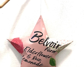 May include: A star-shaped hanging ornament made from a pink Belvoir Farm Elderflower & Rose Lemonade label. The design features black text, a pink rose illustration, and green leaves, suspended by a thin white string from a wooden branch.